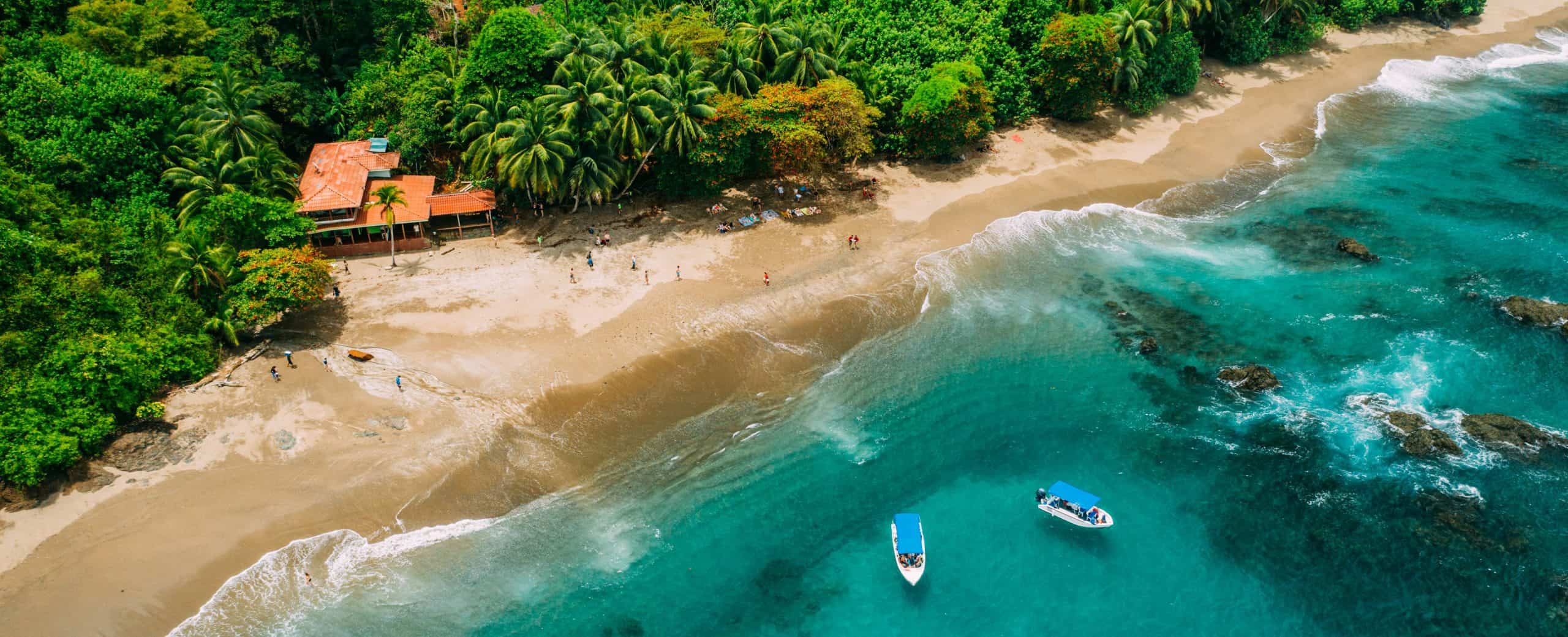 Aerial view of a pristine tropical beach with turquoise waters, lush greenery, and boats anchored ne.