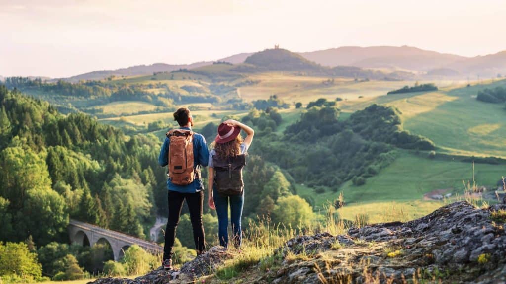 Two women enjoying a scenic countryside view, hiking in lush green hills with a historic bridge in t.