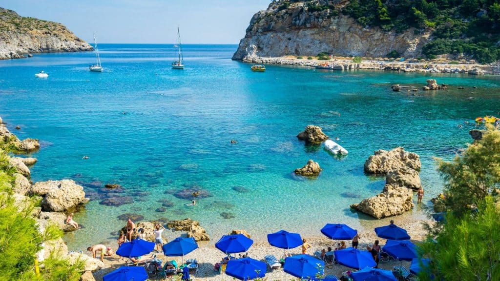 Beautiful seaside view with blue umbrellas, rocky shoreline, and sailing boats in clear waters.