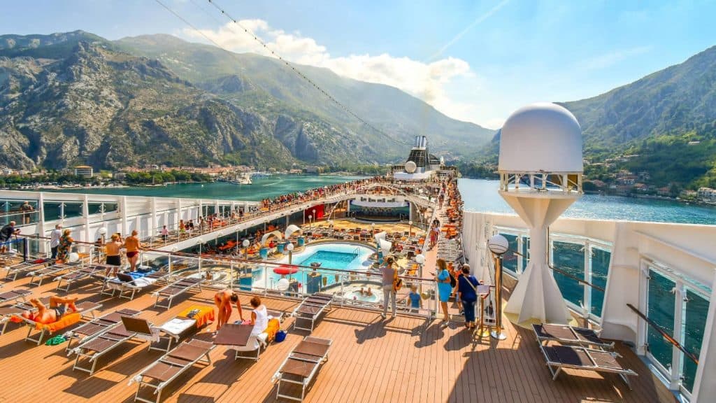 Cruise ship deck with pool, sun loungers, and mountain scenery in the background.